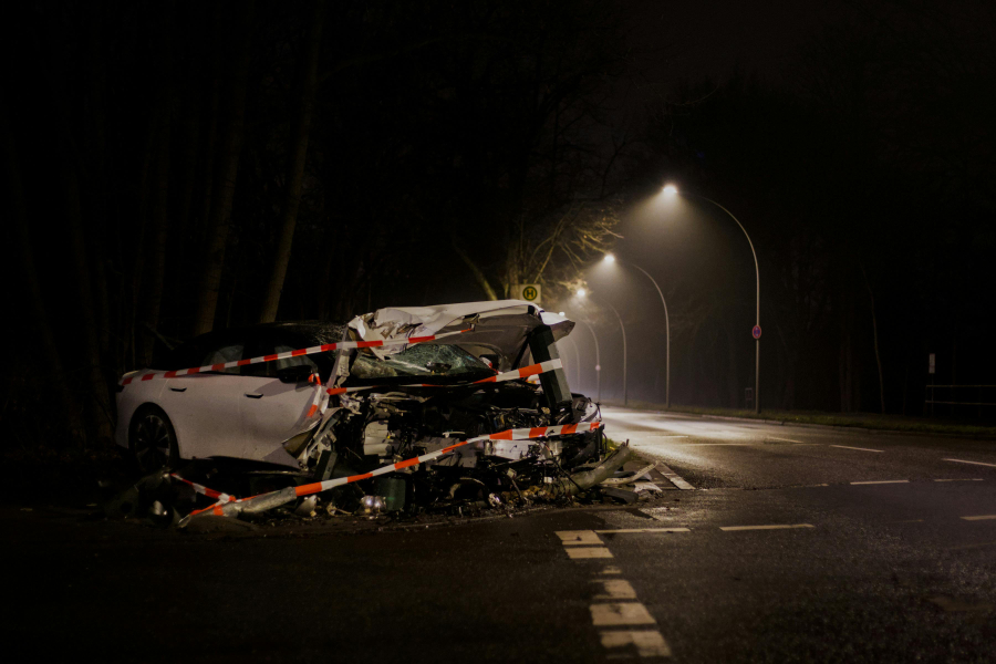 Night scene of a white car wreck on a road, heavily damaged and surrounded by red-and-white caution tape under streetlights at night to indicate a crash site