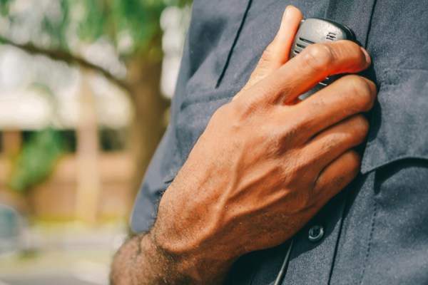 Close-up of a man holding a handheld radio to his chest outdoors; wearing a dark shirt.