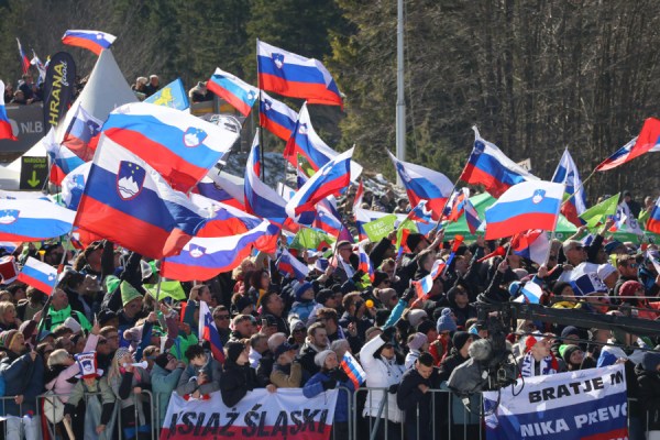 Crowd at an outdoor rally waving Slovenian flags and banners above a barricade area.