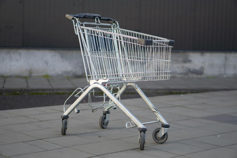 Shopping cart on a paved sidewalk, empty and ready for groceries, with four wheels and a handle.