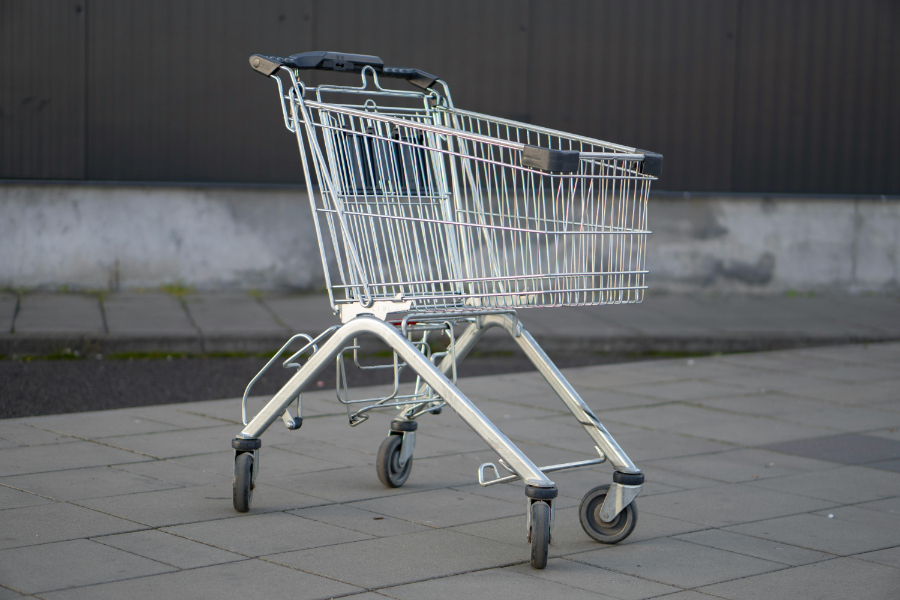 Shopping cart on a paved sidewalk, empty and ready for groceries, with four wheels and a handle.