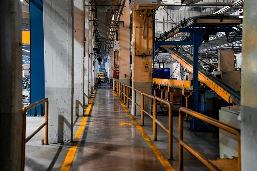 Factory corridor in a manufacturing plant with yellow safety rails and overhead machinery along a narrow walkway.